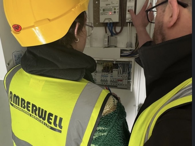 Jess in a hi-vis vest and yellow hard hat looking at some electrics with her mentor