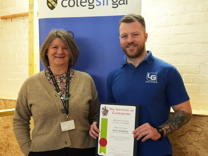 Tom holding his certificate in a carpentry workshop with Helen head of construction