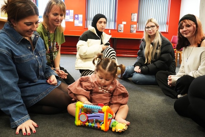 Liwsi with her mum Zoe, some students and tutor watching her playing on the floor, they are all sat on the floow