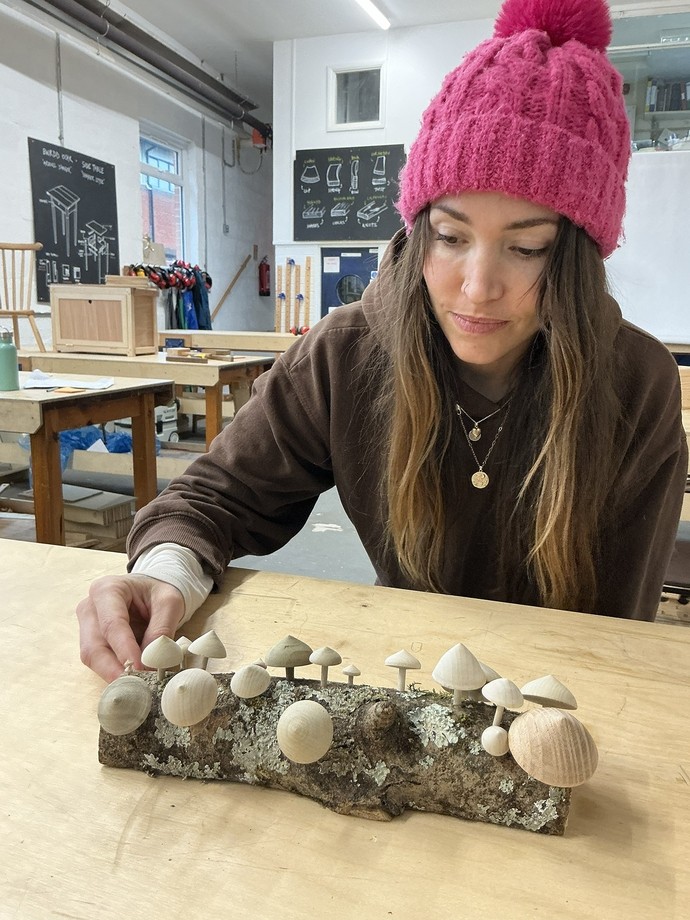 A student in a red bobble hat with her work which is wooden mushrooms on a small log