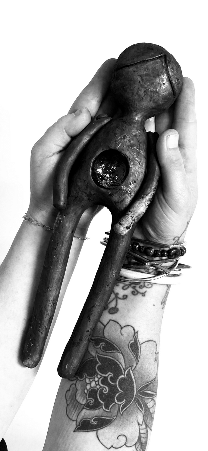two human hands/arms, one with a tattoo. It's all black and white and they are holding a ceramic piece shaped like a simplified human shape
