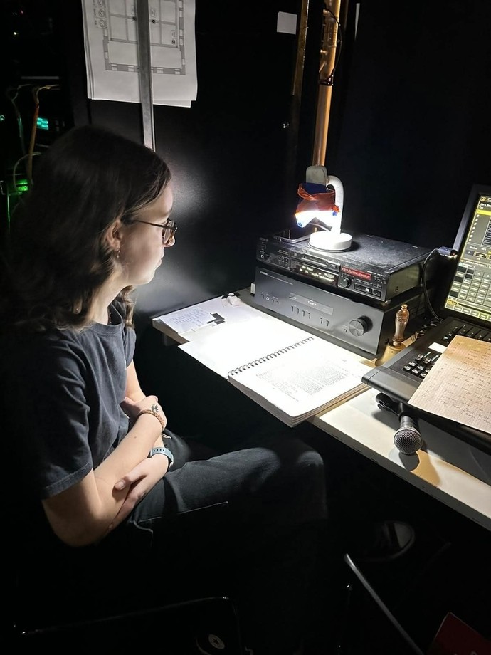 Dulcie sitting in a dimly lit room with headphones on looking at a lit up creative system (a feeling of working behind the scenes)