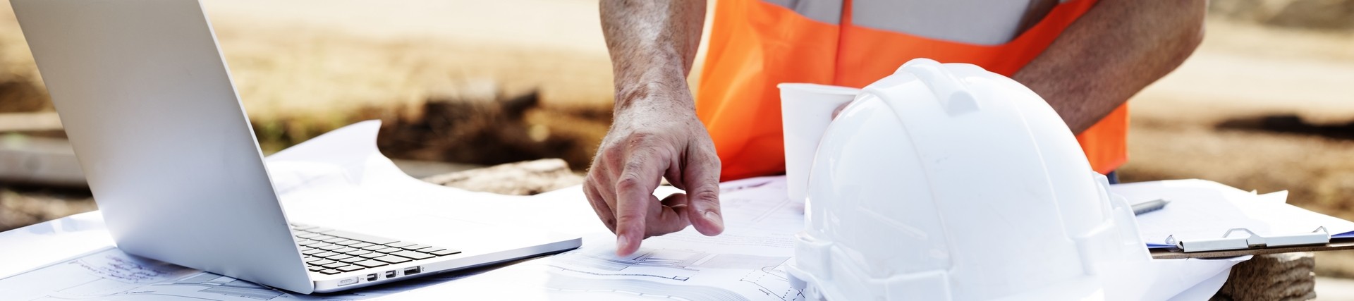 close up shot of a table with building plans being inspected bu a person in an orange high-vis vest. theer's a white hard hat on the table.