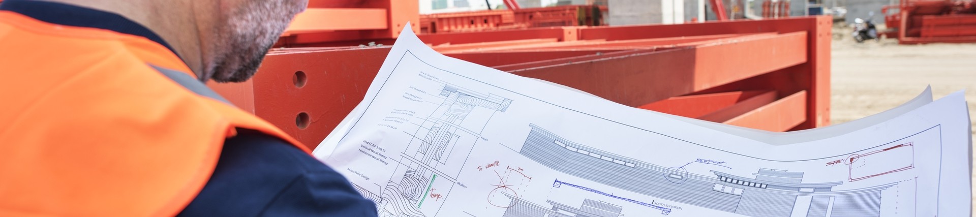 over the shoulder shot of a man in orange high-vis vest and white hard hat looking at building plans
