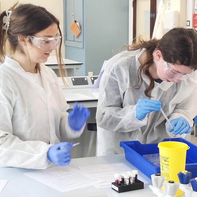 Two students in lab coats and glasses wearing PPE gloves conducting an experiment